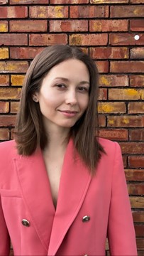 A young female adult with long brown straight hair, wearing a peach coloured blazer/jacket with gold buttons standing against a red brick wall.